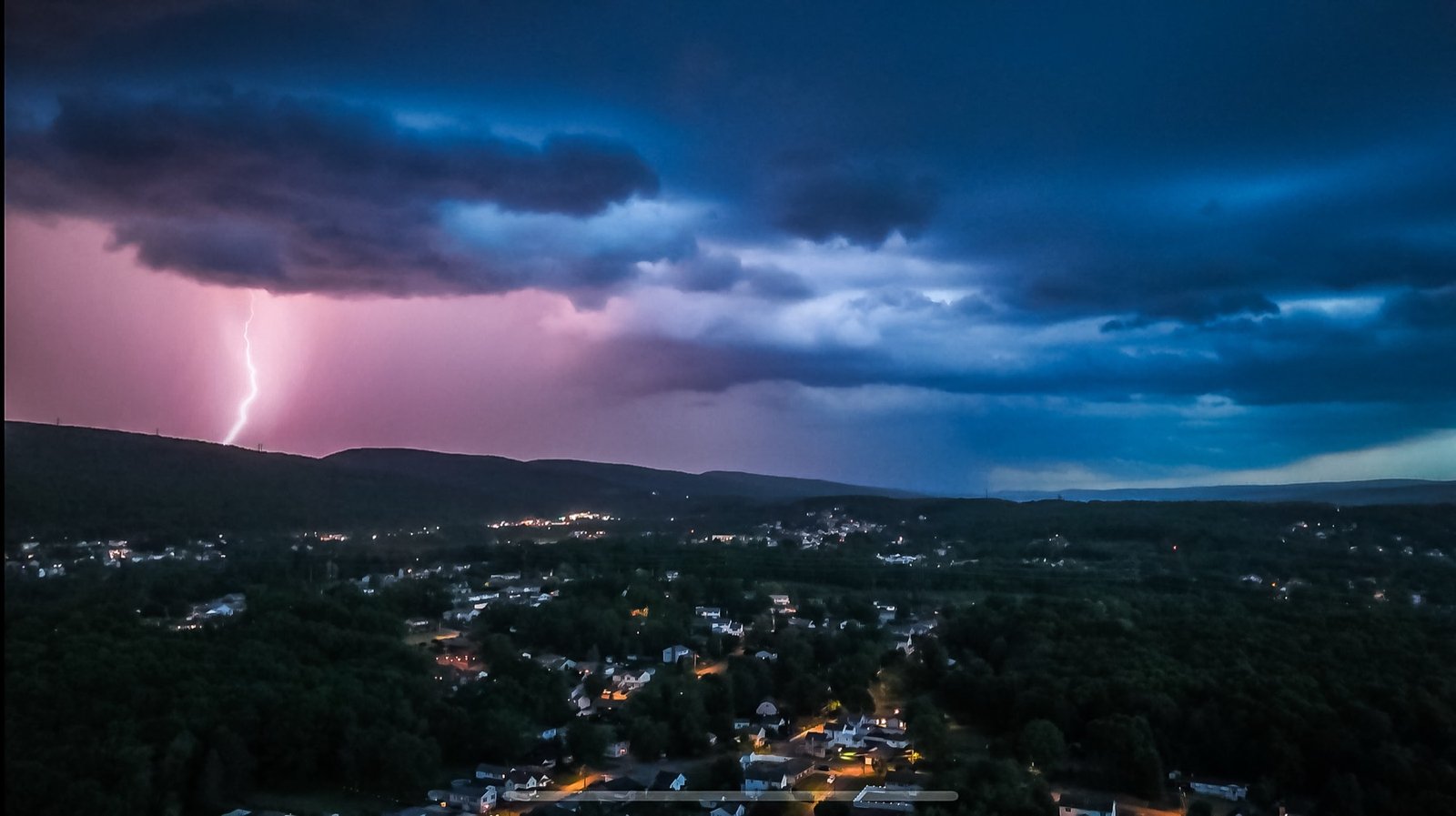 Lightning striking beyond a town at dusk under purple storm clouds